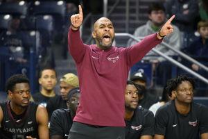 Ein Mann in einem roten Hemd und schwarzen Shorts steht am Spielfeldrand einer Basketballhalle und unterhält sich mit einer Gruppe von Menschen, einige sitzen und einige stehen.