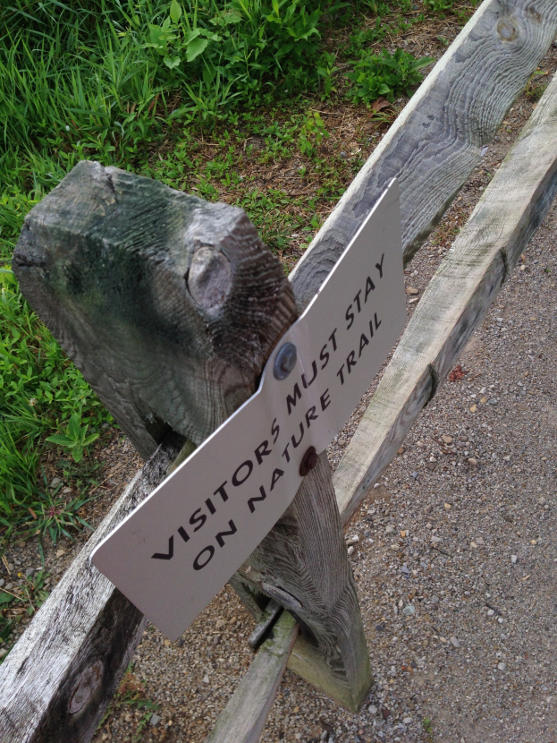 Holzzaun mit einem Schild, auf dem "Besucher sollten auf dem Naturweg bleiben" steht, in üppiger grüner Umgebung mit kleinen Steinen.