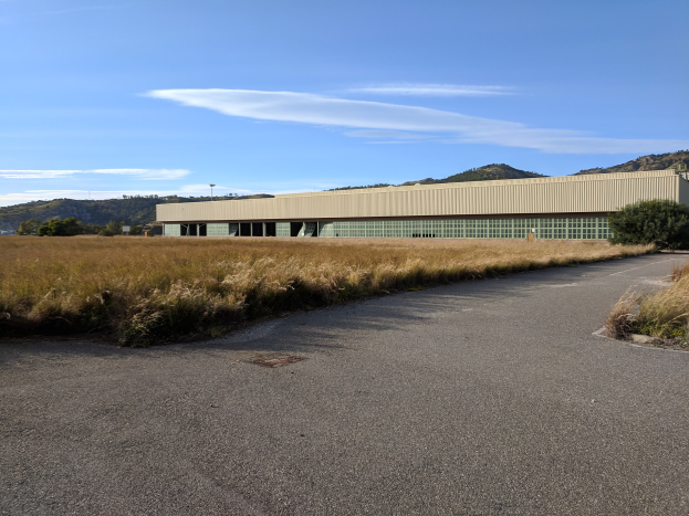 Leere Straße, die zum Hauptquartier des San Francisco International Airport führt, ein großes Gebäude auf einem Feld mit umgebenen Pflanzen und Bäumen, Hügel und bewölkter Himmel im Hintergrund.