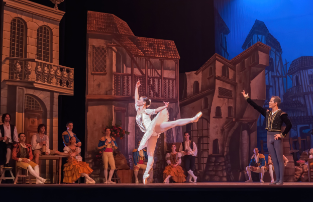 Zwei Tänzer performen auf einer Bühne im Royal Opera House in London, mit einem Publikum im Vordergrund und einer projizierten Stadtlandschaft als Hintergrund.
