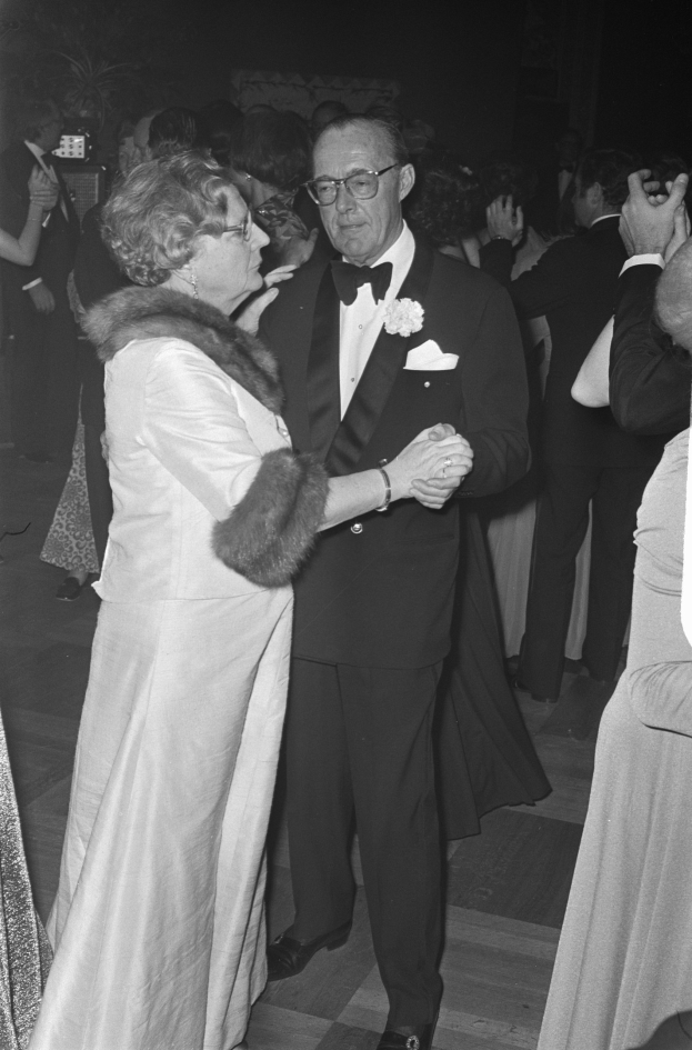 Formal gekleideter Mann und Frau tanzen zusammen auf einer Party, während andere im Hintergrund stehen, einige halten Kameras.