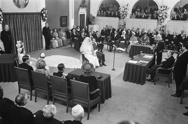 Schwarzes und weißes Foto einer Hochzeitszeremonie in einem großen Raum mit sitzenden und stehenden Gästen, Tischen mit Mikrofonen und Papieren, einer hinteren Wand mit einem gerahmten Foto, Blumenbouquets, einem Vorhang und einem Podium mit einem Mikrofonständer.