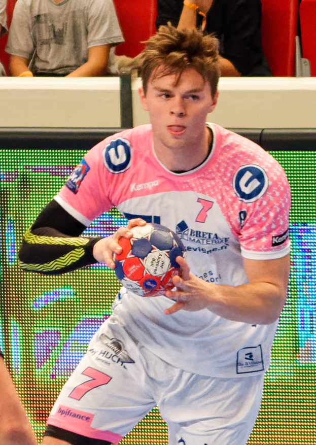 Ein Mann in einem pinken Hemd mit einem Fußball, daneben eine andere Person und im Hintergrund sitzende Menschen.
