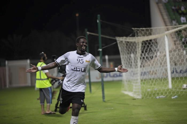 Ein Mann in einem weißen Hemd und schwarzen Shorts rennt auf einem Fußballfeld mit ausgestreckten Armen, umgeben von stehenden Zuschauern, mit einem Torpfosten und sitzendem Publikum im Hintergrund, feiernd nach einem Torerfolg.