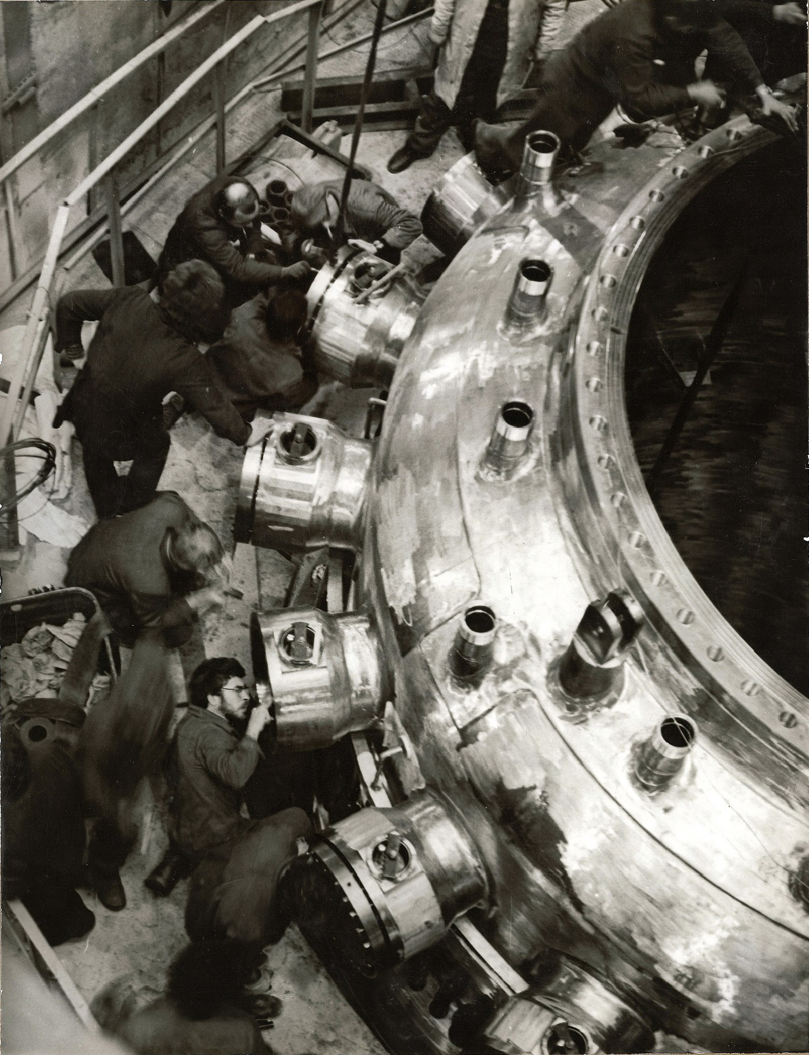 Schwarzes und weißes Bild von Männern, die an einem großen Metallreaktor in einer industriellen Fabrikumgebung arbeiten, mit Treppen, Geländern und einer Wand im Hintergrund.