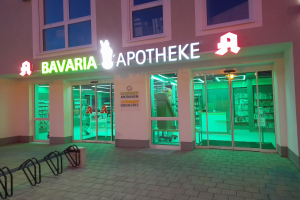 Ein gläsernes Gebäude mit einer beleuchteten Schilder, auf dem "Bavaria Apotheke" steht, das Regale mit verschiedenen Gegenständen zeigt und einige Gegenstände auf der linken Seite des Eingangs verstreut sind.