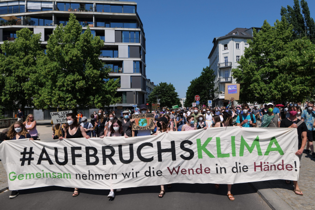Eine Gruppe von Menschen mit Masken hält ein Banner mit der Aufschrift 'Aufbruchsklima' vor einem Gebäude, umgeben von Bäumen und einem klaren Himmel.