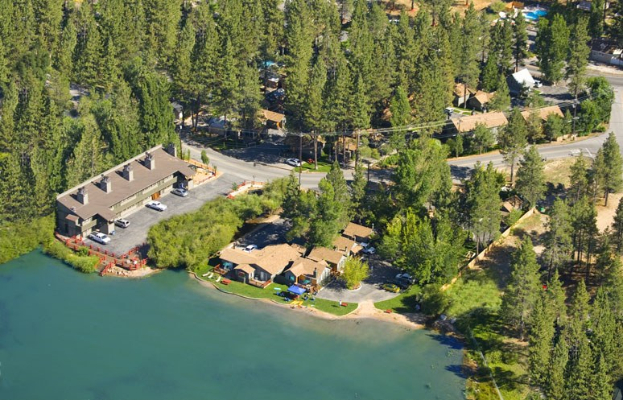 Luftaufnahme eines Resorts am Seeufer, umgeben von Bäumen, Häusern, Fahrzeugen, Straßen, Masten und Gras, mit dem See unten.