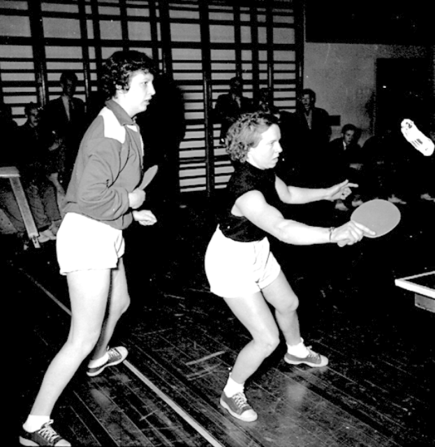 Zwei Frauen beim Tischtennis auf einem Court, jede hält einen Schläger in der Hand, mit einigen Zuschauern im Hintergrund und einem Tischtennis-Tisch auf der rechten Seite, in Schwarz-Weiß.