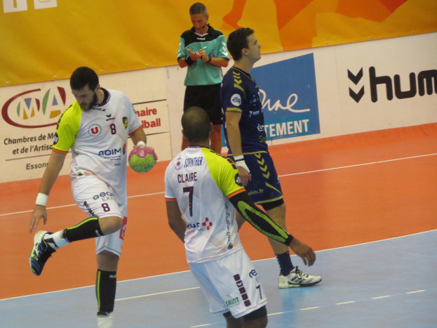 Eine Gruppe von Männern, die Handball auf einem Platz spielen, wobei einer einen Ball hält und im Hintergrund Schilder mit Text zu sehen sind.