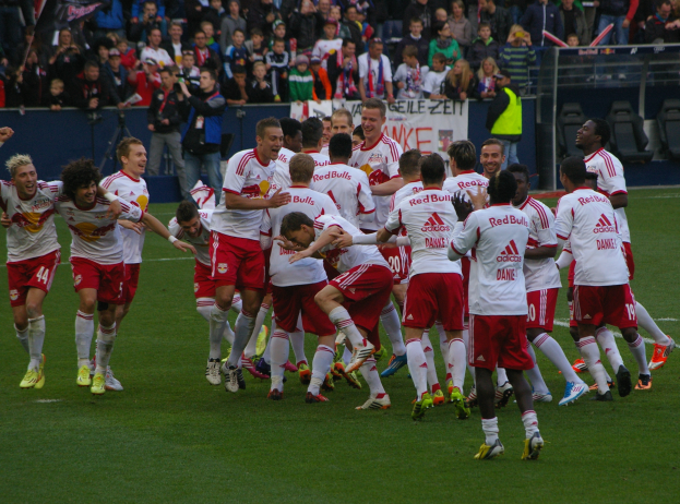 Eine Gruppe von Fussballspielern feiert ein Tor auf einem grünen Feld, mit Zuschauern in den Rängen, die Kameras und Banner halten, und Geländern und Stühlen um das Feld herum.