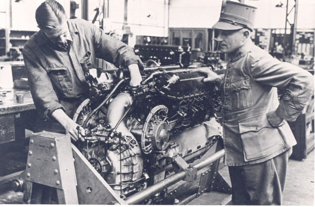Zwei Männer in einem Schwarz-Weiß-Bild arbeiten an einem Flugzeugturbinen-Triebwerk in einer Fabrik, mit einigen anderen Personen im Hintergrund.