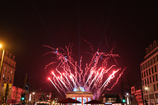 Eine belebte Stadtstraße in Berlin an Silvester, voller Menschen, Fahrzeuge und Gebäude, erleuchtet von Feuerwerk und Gebäudelichtern, schafft eine festliche Atmosphäre.