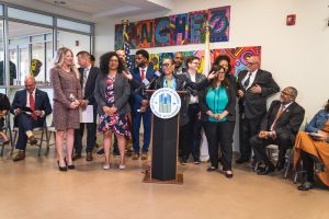 Gruppe von Menschen vor einem Rednerpult mit Mikrofon und einer Tafel mit der Aufschrift "Sancho-Stadtrat tritt dem Stadtrat bei", mit sitzenden und stehenden Teilnehmern, Fahnen, Feuerlöscher und Deckenbeleuchtung.