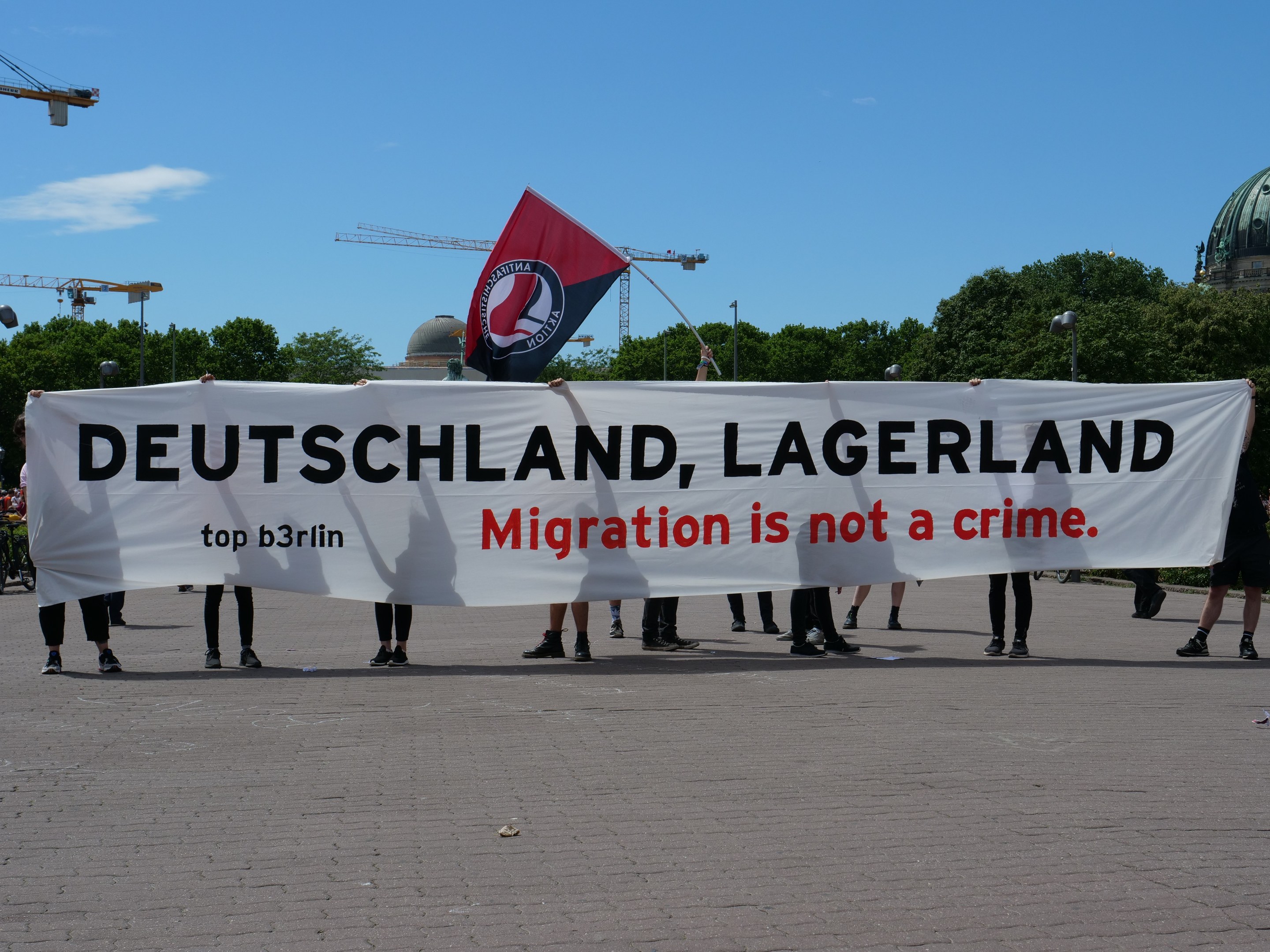Eine Gruppe von Menschen hält ein Transparent mit der Aufschrift "Deutschland, Lagerland Migration ist kein Verbrechen" und eine Flagge, im Hintergrund sind Straßeninfrastruktur, Bäume, ein Gebäude und ein bewölkter Himmel zu sehen.