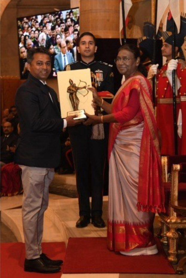 Ein Mann in einem schwarzen Blazer, weißem Hemd und grauen Hosen, der eine Trophäe hält, neben einer Frau in einem aschfarbenen Sari auf einem roten Teppich bei einer Preisverleihung, mit Menschen, Stühlen, Fahnen und einem Bildschirm im Hintergrund.