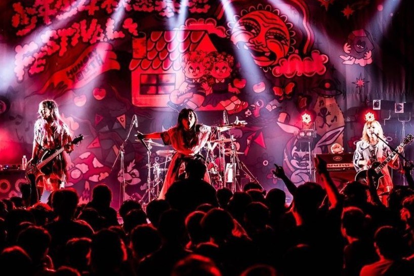 Eine Live-Aufführung einer japanischen Rockband auf der Bühne mit Musikern, die Gitarren spielen und singen, Mikrofone und Ständer vorne, eine beleuchtete Wandmalerei und ein sichtbares Publikum unten.