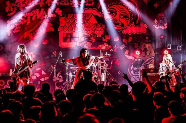 Eine Live-Aufführung einer japanischen Rockband auf der Bühne mit Musikern, die Gitarren spielen und singen, Mikrofone und Ständer vorne, eine beleuchtete Wandmalerei und ein sichtbares Publikum unten.