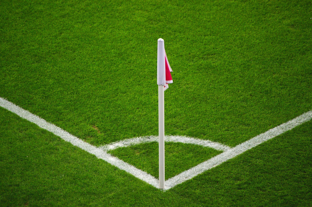 Ein Fussballfeld mit gruener Gras, weissen Begrenzungslinien und einem weissen Pfahl mit einer rot-weissen Flagge, die den Elfmeterpunkt markiert.