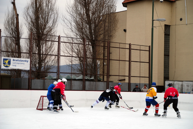 Menschen beim Eisschlittschuhlaufen auf einer Eisbahn mit Gebäuden, Bäumen, einer Straßenlaterne, einem Namensschild und Zäunen im Hintergrund unter einem Himmel.