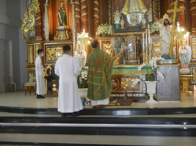 Drei Priester vor einem Altar mit Statuen, Kerzen, Blumenvasen und anderen dekorativen Gegenständen in einer Kirche.