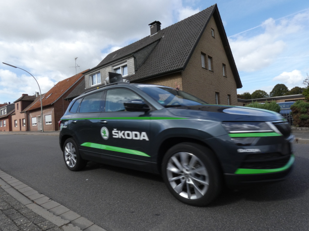 Schwarzer und grüner Skoda Kodiaq-Elektro-SUV auf einer Stadtstraße mit Gebäuden, Bäumen und bewölktem Himmel im Hintergrund.