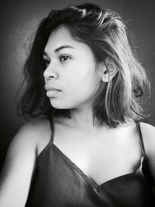 Eine selbstbewusste Frau in einem schwarzen Kleid posiert in einem Schwarz-Weiß-Foto, ihr glatter Bob und das natürliche Make-up unterstreichen ihre selbstbewusste Haltung vor einer neutralen Wand.