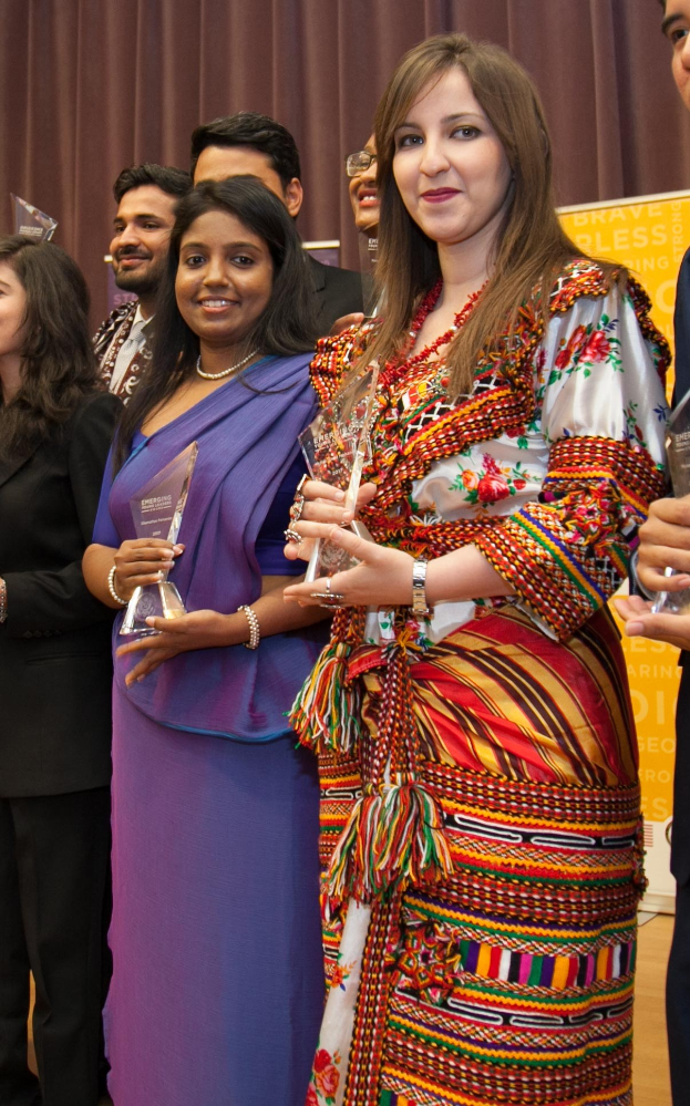 Eine Gruppe von Menschen auf einer Bühne mit einer Frau in der Mitte, die eine Trophäe hält, vor einem Vorhang und einer Tafel mit der Aufschrift "Bravo Segen Award Gewinner".