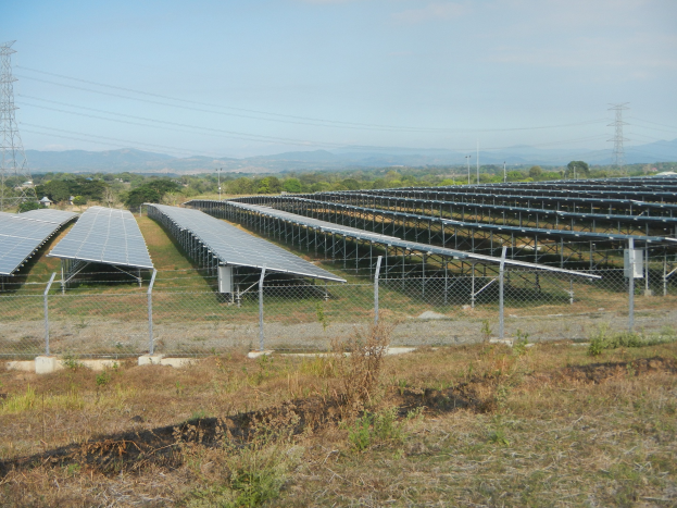 Luftaufnahme eines Solarpanelfelds mit einem Zaun im Vordergrund, Gras, Pflanzen, Bäumen, Übertragungsmasten mit Drähten, Hügeln und einem klaren blauen Himmel.