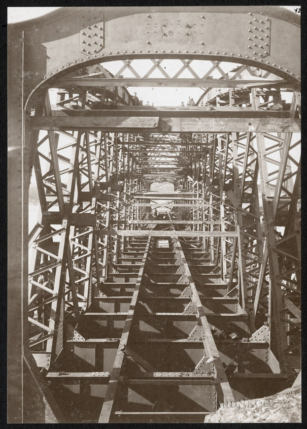 Schwarz-weiß-Foto einer alten Brücke mit Metallstangen und Treppen, die hinaufführen, mit Text unten.