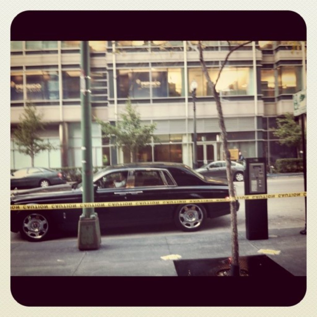 Ein Auto ist in der Mitte des Bildes mit Pfählen und Bäumen drumherum, ein Gebäude und weitere Fahrzeuge im Hintergrund und ein Gehweg unten.