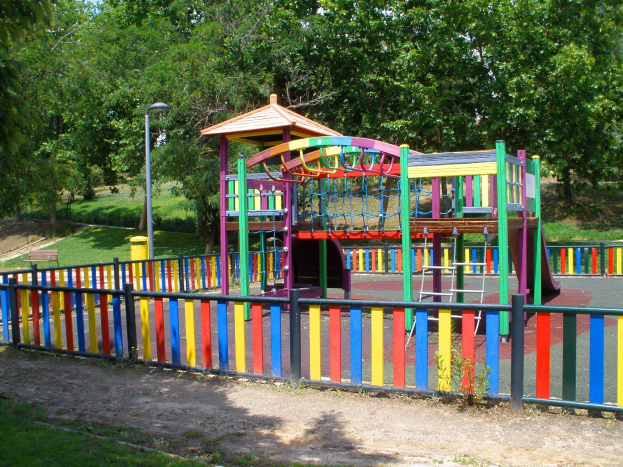 Ein Spielplatz mit farbenfroher Spielstruktur, umgeben von einem Zaun, Pfosten, Lichtern, B├Ąnken, Gras, Pflanzen und B├Ąumen unter einem klaren blauen Himmel.
