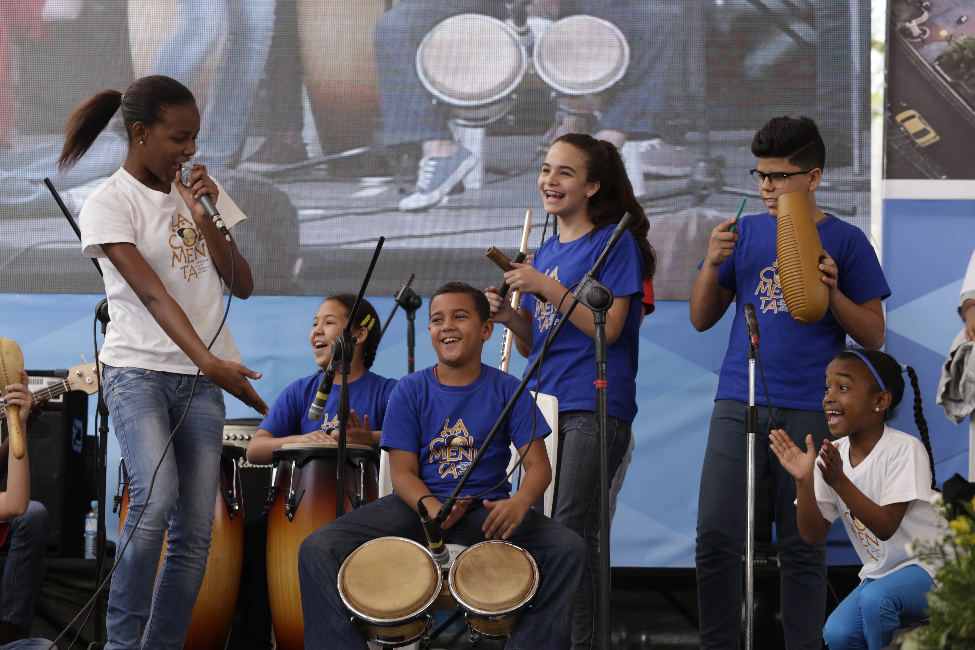 Eine Gruppe von Kindern, die auf einer Bühne Musikinstrumente spielen, mit einer Frau, die ein Mikrofon hält, umgeben von Mikrofonständern, einem Banner im Hintergrund und Pflanzen auf der rechten Seite.