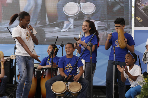 Eine Gruppe von Kindern, die auf einer Bühne Musikinstrumente spielen, mit einer Frau, die ein Mikrofon hält, umgeben von Mikrofonständern, einem Banner im Hintergrund und Pflanzen auf der rechten Seite.