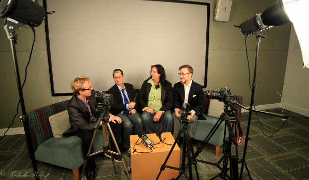 Eine Gruppe von drei Personen sitzt auf einem Sofa und gibt ein Interview, wobei eine Person links in einem schwarzen Mantel und Hose, eine Person in der Mitte mit einem Mikrofon und einer blauen Krawatte und eine Person rechts mit einem Lächeln zu sehen ist. Ein Mikrofonrekorder steht in der Mitte, Kameras sind auf der rechten und linken Seite und große Scheinwerfer sind vorhanden, sowie ein Projektionsschirm im Hintergrund.