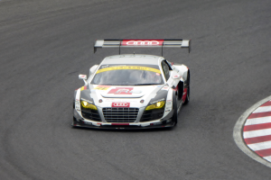 Ein leuchtend rotes und weißes Audi R8 LMS GT3-Rennfahrzeug mit dem Audi-Logo auf der Seite, gefahren von einer Person in einem schwarzen Rennanzug und Helm, fährt um eine Rennstrecke.