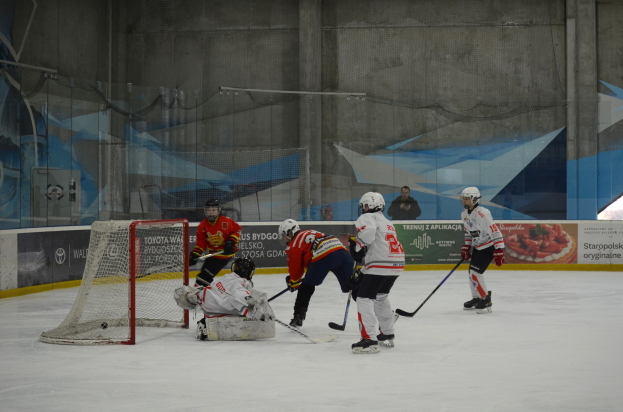 Gruppe von Menschen, die Eisockey auf einem Eisplatz spielen, tragen Helme und halten Eishockeyschläger, mit einem Tor auf der linken Seite und Bannern im Hintergrund.