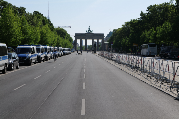 Lange Reihe von Polizeiwagen, die entlang einer Straße vor dem Reichstagsgebäude in Berlin geparkt sind, mit Fahrradfahrern und Fußgängern, Absperrungen, Bäumen und dem Torbogen mit Statuen im Hintergrund.