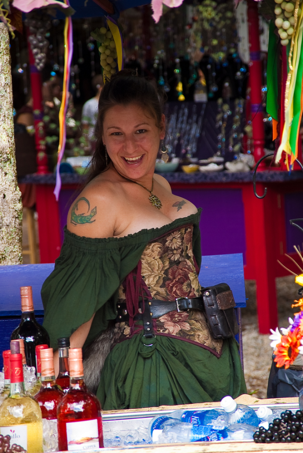 Eine Frau in einem grünen Kleid lächelt und zeigt ein Tattoo auf ihrer Hand, sitzt an einem Tisch mit Flaschen, Blumen, Früchten und verschiedenen Gegenständen im Hintergrund.