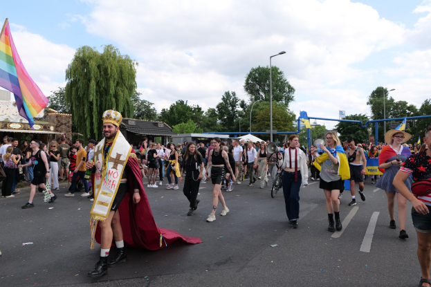 Eine Gruppe von Menschen marschiert bei der Gay Pride Parade 2018 mit einer Regenbogenflagge und Musikinstrumenten, einige tragen Mützen, vor einem Hintergrund aus Laternenpfählen, Bäumen, Hütten und einem bewölkten Himmel.