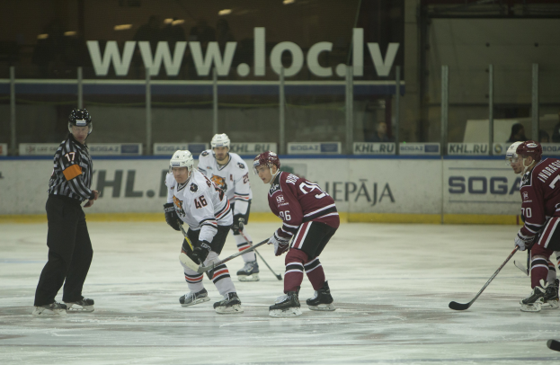 Gruppe von Menschen, die Eisstockschießen auf einem Eisplatz betreiben, mit Helmen und Eishockeyschlägern, mit einem Schiedsrichter auf der linken Seite und Zuschauern im Hintergrund.