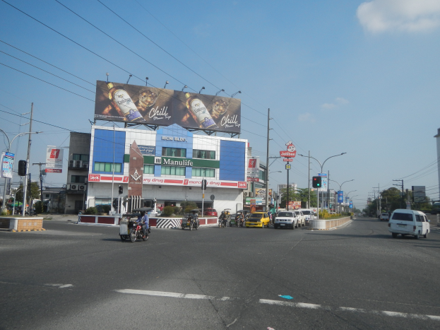 Eine belebte Stadtstraße mit starkem Verkehr, hohen Gebäuden, Strommasten, Drähten, Laternen, Verkehrsampeln und verschiedenen Schildern, mit einem Plakat in der Mitte, Bäumen und einem bewölkten Himmel im Hintergrund.