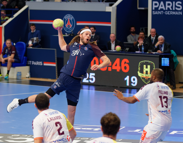 Eine Gruppe von Männern spielt Handball auf einem Platz mit einem Ball in der Luft, Zuschauern im Hintergrund und einem Tisch mit einem Monitor, Flaschen und anderen Gegenständen, mit einer Tafel, die Text und Logos anzeigt, die auf ein Spiel zwischen Paris Saint-Germain und Olympique Lyon hinweisen.