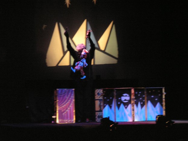 Eine Person auf einer Bühne, die mit Puppen spielt und die Fäden hält, mit Dekoration und Lichtern im Hintergrund.