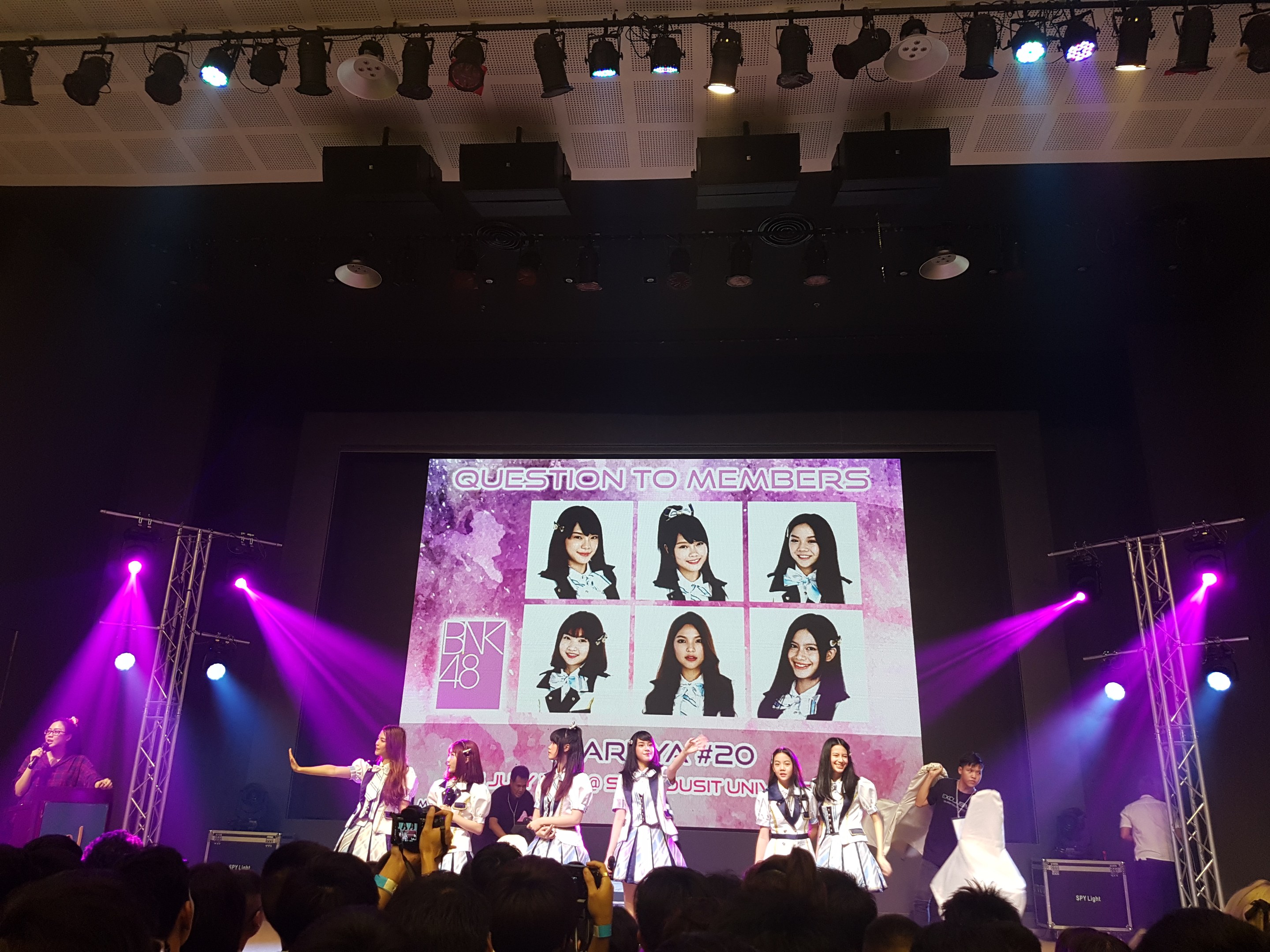Eine Gruppe von Menschen, die auf einer hell erleuchteten Bühne mit einem großen Bildschirm, der BK48-Mitglieder zeigt, performen, während das Publikum darunter aufnimmt.