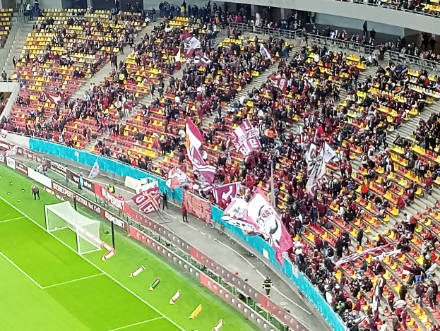 Ein großes Stadion voller Zuschauer bei einem Fußballspiel mit einem Tor und Netz auf der linken Seite, einigen Menschen, die stehen und auf Stühlen sitzen, mehreren Tafeln mit Text und einer Treppe, Geländer und einer Wand im Hintergrund.