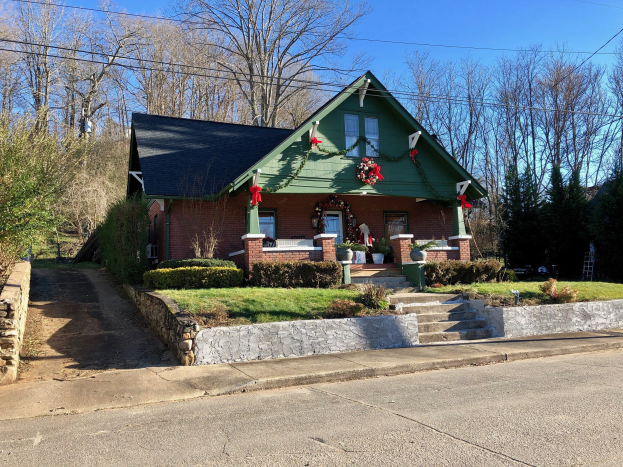 Ein grünes Haus mit einem Kranz an der Vorderseite, umgeben von Pflanzen, Gras, Treppen, einer Wand, Bäumen, Drähten und einem bewölkten Himmel in einer städtischen Umgebung, verfügbar zur Miete.