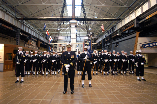 Eine Gruppe uniformierter Männer mit Mützen und Gewehren steht in einer großen Halle mit Flaggen, Wandtafeln und Deckenleuchten und -stangen.