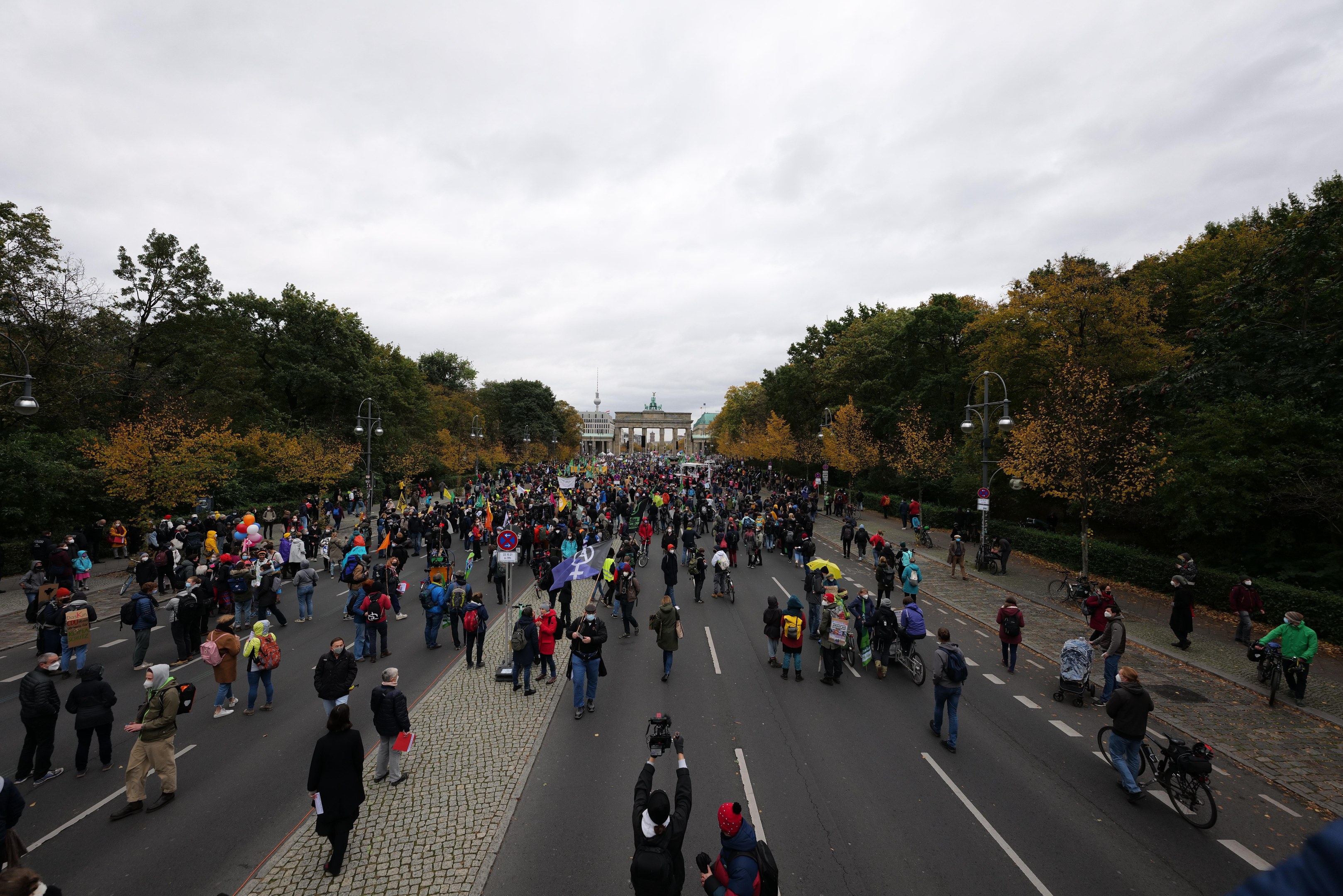 Eine große Gruppe von Menschen marschiert auf einer von Bäumen gesäumten Straße mit Laternenpfählen, hält Kameras und nimmt an einer Protestdemonstration in Berlin teil, mit einem Gebäude und einem klaren Himmel im Hintergrund.