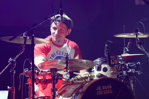 Ein Mann in einem roten und weißen T-Shirt spielt Schlagzeug auf der Bühne während eines Konzerts, er strahlt umgeben von Mikrofonen.